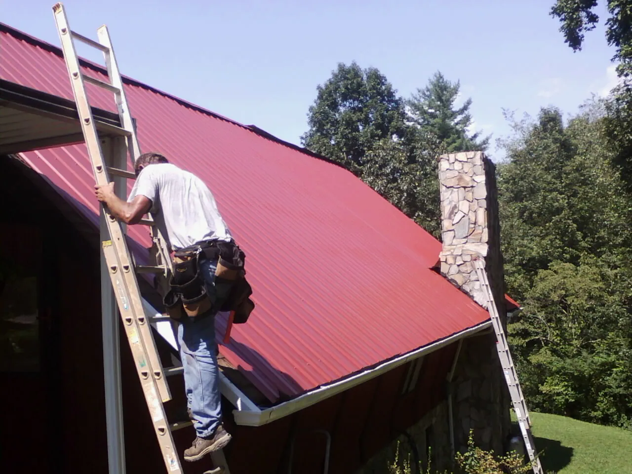 Roofer on a ladder beside a red metal roof and stone chimney — Soffit & Fascia Repair by Roofing Brothers in North Adams
